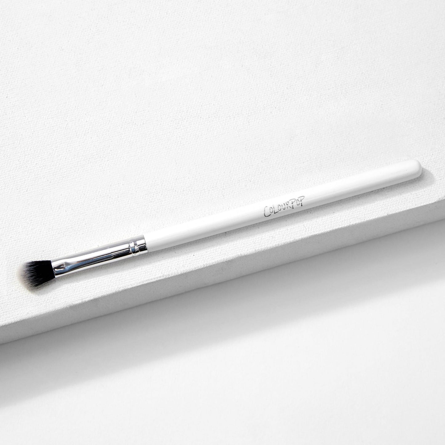 ColourPop Blending Brush makeup brush resting diagonally on a white textured surface, featuring fluffy angled black-to-white bristles, a silver ferrule, and a slim white handle, made for seamless eyeshadow blending.